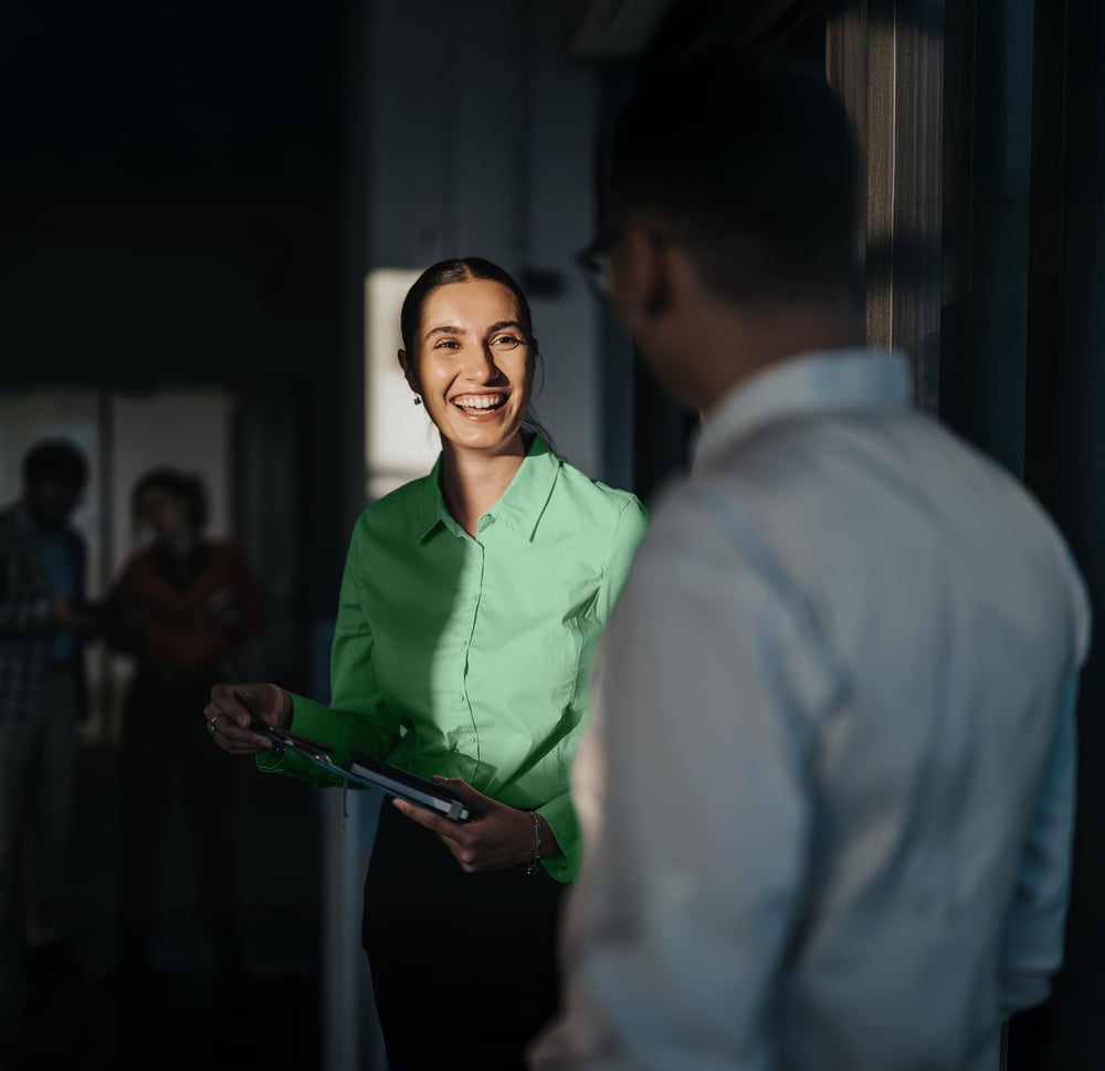 A smiling woman in a green blouse holding a tablet, talking to a colleague in an office hallway.