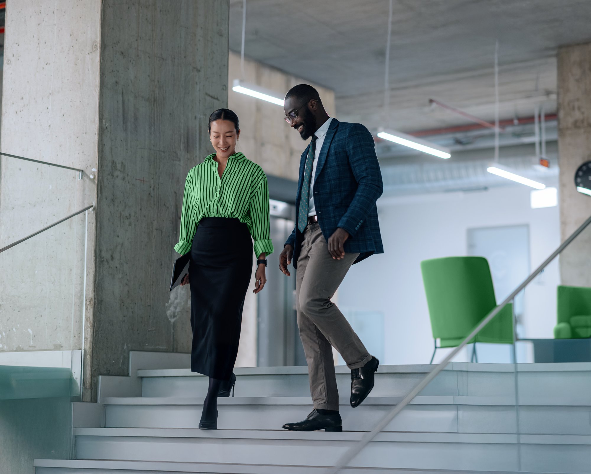 Two colleagues, a woman in a green striped blouse and a man in a suit, walking down the stairs together in a modern office building.