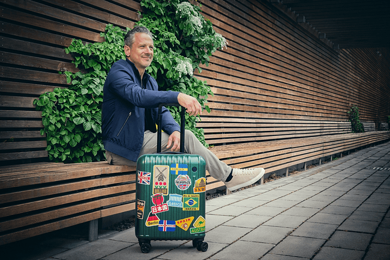 Mann sitzt entspannt auf einer Holzbank vor einer begrünten Wand und hält den Griff eines grünen Rollkoffers mit bunten Reiseaufklebern aus verschiedenen Ländern