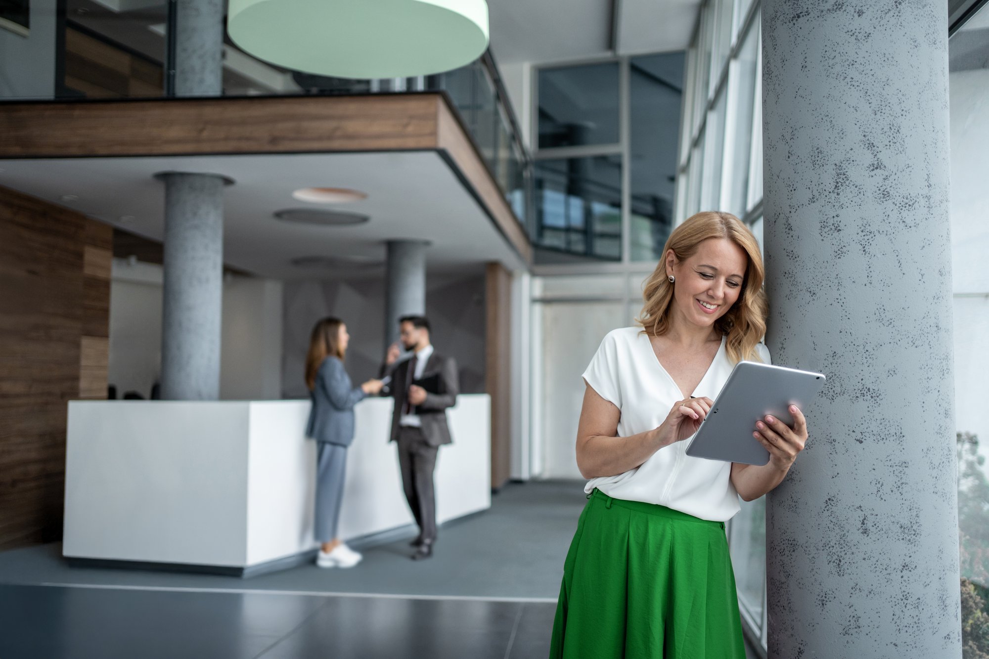A woman in a white blouse and green skirt leaning against a pillar, smiling while using a tablet in a bright office lobby. A woman in a white blouse and green skirt leaning against a pillar, smiling while using a tablet in a bright office lobby.