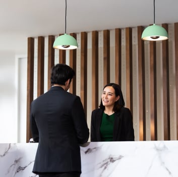 Rezeptionistin hinter einem Marmortresen lächelt einen Gast im Anzug an; über ihnen hängen zwei grüne Pendelleuchten, im Hintergrund eine Wand mit vertikalen Holzleisten.