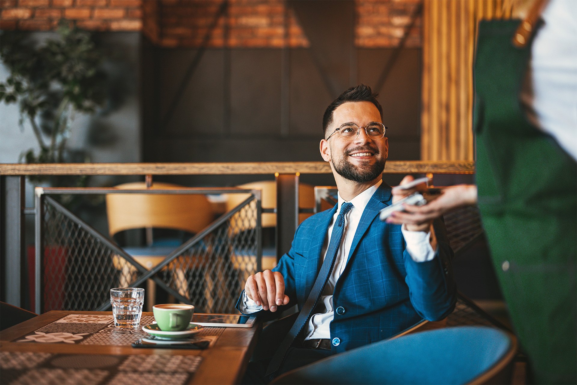 A smiling businessman in a suit uses his smartphone for a contactless payment in a cozy cafeteria setting.